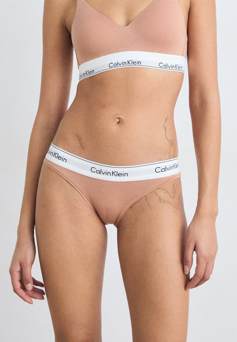 Sutien în culoarea nud de la Calvin Klein și bikini asortați, cu un brâu alb ce poartă un logo. Material neted, design minimalist, talie medie.