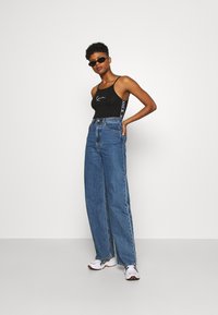 Svart tettsittende singlet med hvitt logo, kombinert med høyt liv og vide blå denimjeans. Brukt med hvite joggesko og solbriller.