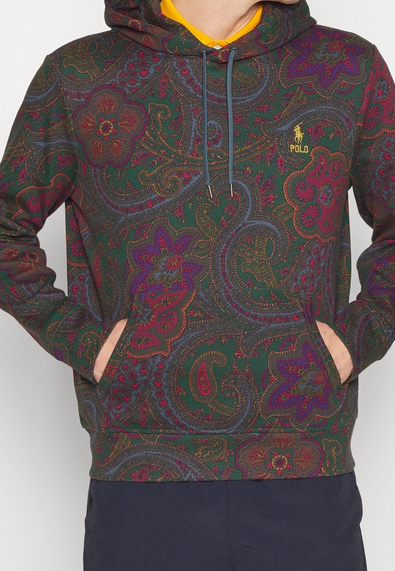 Personne portant un sweat à capuche à motif cachemire multicolore avec les mains dans la poche avant et un t-shirt jaune visible au niveau du cou.
