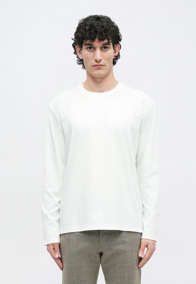 Weißes Langarmshirt mit rundem Ausschnitt, aus einem glatten Stoff gefertigt, kombiniert mit hellgrauen, strukturierten Hosen. Einfaches, minimalistisches Design.