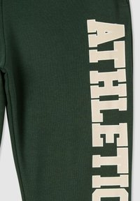 Zielone spodnie dresowe wykonane z fakturowanej tkaniny, z dużym kremowym napisem "ATHLETIC" ułożonym pionowo po lewej stronie.