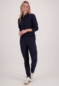 Lachende vrouw met lang blond haar, gekleed in een marineblauw overhemd met knopen, bijpassende broek en witte sneakers met zwarte strepen.