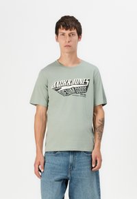 Grøn bomulds t-shirt med rund hals; har et iøjnefaldende sort-hvid grafisk print på fronten. Kombineret med blå denim jeans.