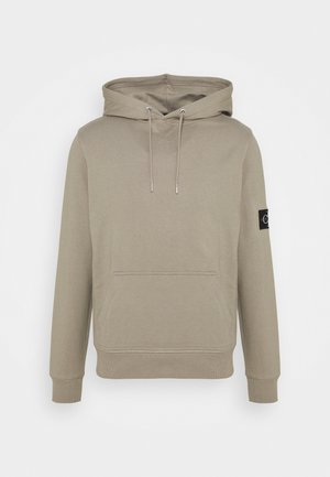 Sweat à capuche beige, avec poche kangourou à l’avant, capuche avec cordon, poignets et ourlet côtelés, et un patch logo noir sur la manche gauche.
