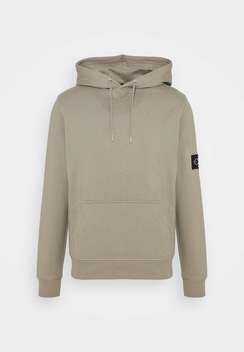 Sweat à capuche beige, avec poche kangourou à l’avant, capuche avec cordon, poignets et ourlet côtelés, et un patch logo noir sur la manche gauche.