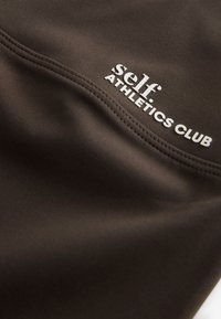 Close-up of dark fabric with stitched seam and white text reading "self ATHLETICS CLUB" on the surface.
