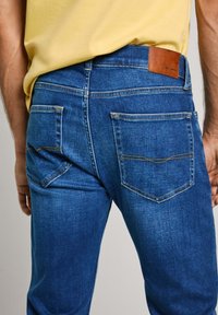 Jean en denim avec un lavage bleu foncé, présentant une écusson en cuir sur la taille, des surpiqûres jaunes et deux poches arrière.