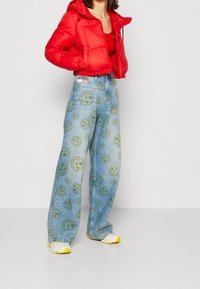 Giacca a piumino rossa con cappuccio, abbinata a jeans a gamba larga blu con motivo a faccina sorridente. Sneakers con dettagli gialli completano il look.