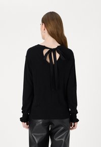 Schwarzer Pullover mit Rundhalsausschnitt, Schlüsselloch-Rücken und Kordel-Detail. Lange Ärmel mit Rüschen am Saum. Kombiniert mit glänzenden schwarzen Hosen.