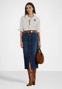 Camicia bianca con bottoni, logo blu navy; maglietta a righe sotto, gonna a matita in denim con spacco anteriore; cintura marrone; stivali alti marroni; borsa in suede marrone.