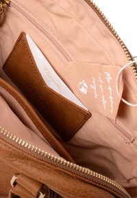 Interior de bolso de cuero marrón con un forro de tela color caramelo, que cuenta con un bolsillo para tarjetas y un pequeño bolsillo con cremallera. Etiqueta notable adjunta en el interior.