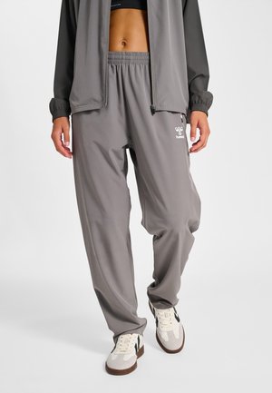 Persoon draagt grijze trainingsbroek, bijpassend grijs jack met rits, zwarte sportbeha en witte sneakers met zwarte details.