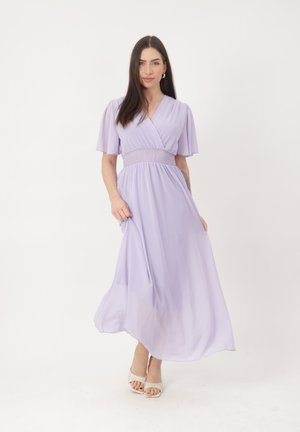 Mujer de pie que lleva un vestido midi fluido de color lila claro con mangas cortas abullonadas y cintura fruncida, combinado con tacones blancos de punta abierta.