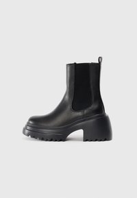 Bottines en cuir noir avec des panneaux élastiques sur les côtés, semelle épaisse et chunky, et détails de texture sur la semelle extérieure. Design minimaliste.