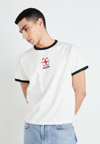 T-shirt blanc avec col et ourlets de manches noirs. Présente un design graphique rouge sur la poitrine. Le matériau semble lisse et léger.