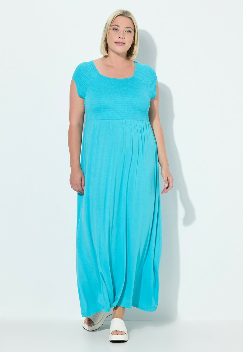 Türkises Maxikleid mit kurzen Ärmeln, strukturiertem Oberteil und fließendem Rock. Kombiniert mit weißen Sandalen mit offener Zehe vor einem hellen Hintergrund.