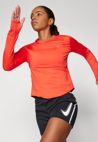 Helles orange langärmliges Sportoberteil mit Daumenlöchern und abgerundetem Saum, kombiniert mit schwarzen Shorts, die an der Seite ein weißes Nike-Logo tragen.
