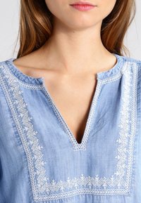 Femme portant un haut bleu clair avec une broderie géométrique blanche autour d'un décolleté à encolure en V, visible des épaules aux lèvres.