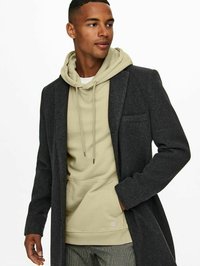 Donkergrijze overjas gemaakt van gestructureerde stof, gelaagd over een lichtbeige hoodie met trekkoorden. Model poseert en toont de outfit.