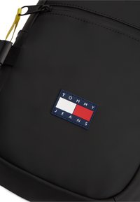 Μαύρο σακίδιο με λεία υφή, με πολύχρωμο λογότυπο σε κόκκινο, λευκό και μπλε χρώμα με το κείμενο "TOMMY JEANS".