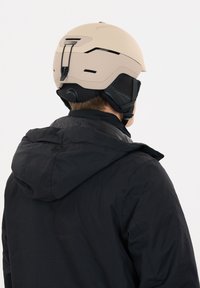 Beigefarbener Skihelm mit Belüftungslöchern und verstellbarem Riemen, getragen von einer Person in einer schwarzen Jacke, von hinten gesehen.
