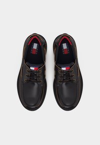 Μαύρα δερμάτινα παπούτσια τύπου boat με καφέ ραφές, μαύρα κορδόνια και το λογότυπο Tommy Jeans στη γλώσσα και στις εσωτερικές σόλες, εμφανιζόμενα από πάνω σε λευκό φόντο.