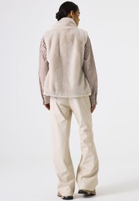Chaleco de piel de color beige con cuello alto, usado sobre una camisa a rayas. Combinado con pantalones de pierna ancha color crema y zapatos oscuros. Diseño minimalista.
