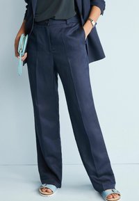 Pantalons larges marine en tissu léger, avec plis sur le devant et poches latérales, associés à des sandales bleu clair et une pochette assortie.