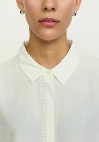 Bijela košulja s ovratnikom i kopčanjem koje sadrži ukrasne gumbe presvučene tkaninom, izrađena od laganog, teksturiranog materijala.