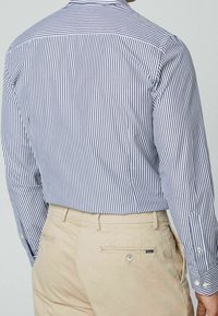 Uomo che indossa una camicia a righe verticali blu e bianche infilata nei pantaloni beige, mostrato di schiena.