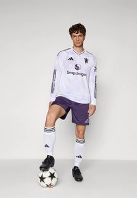 Valkoinen pitkähihaisen pelipaidan, jossa mustia yksityiskohtia ja logo, violettit shortsit, valkoiset sukat mustilla raidoilla ja mustat jalkapallokengät, seisoo pallon päällä.