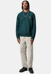 Sudadera verde oscuro con cuello redondo y puños acanalados, que presenta un pequeño logo en dorado. Combinada con pantalones de mezclilla beige claros y zapatos negros.