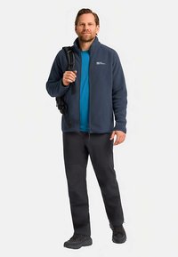 Giacca in pile blu navy con cerniera, abbinata a una camicia blu, pantaloni neri e scarponi da trekking. Zaino sulla spalla, in piedi contro uno sfondo bianco.