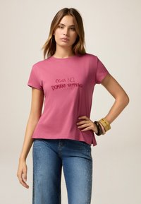 Camiseta de algodón rosa de manga corta que presenta el texto bordado "OGGI NO. DOMANI NEMMENO." Combinada con jeans azules adornados con tachuelas.