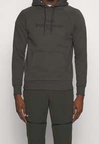 Uomo che indossa un hoodie verde scuro con la scritta "Peak Performance" e pantaloni abbinati con dettagli neri con cerniera sulle ginocchia.
