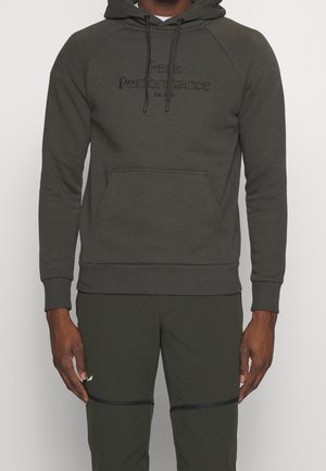 Homme portant un hoodie vert foncé avec le texte "Peak Performance" et un pantalon assorti avec des détails de fermeture éclair noirs sur les genoux.