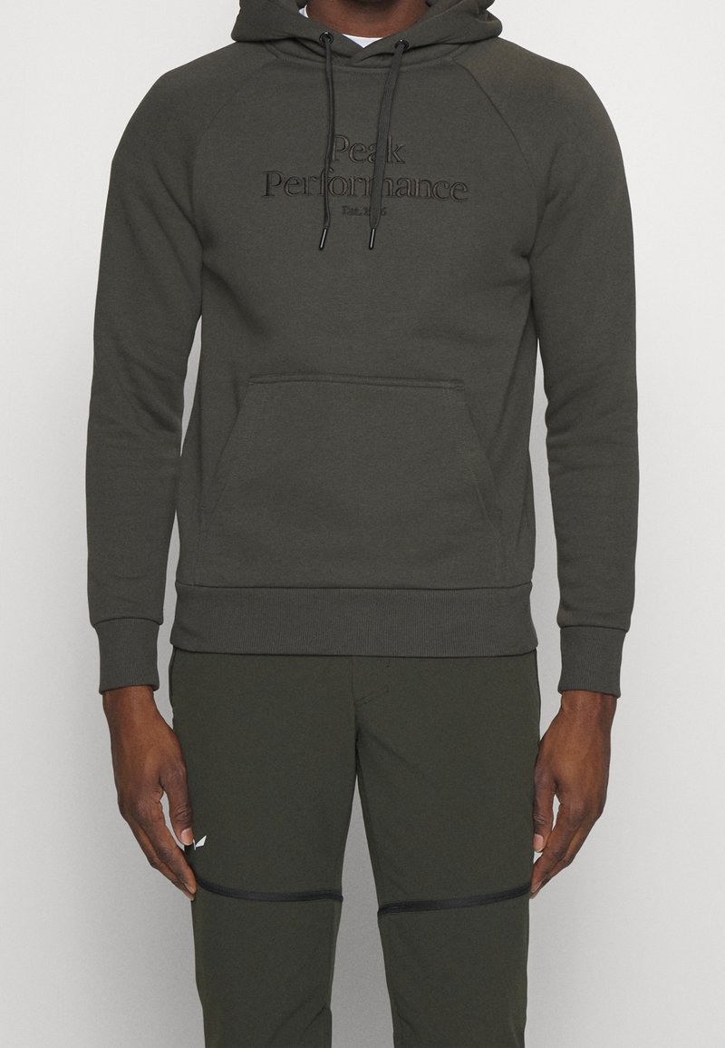 Uomo che indossa un hoodie verde scuro con la scritta "Peak Performance" e pantaloni abbinati con dettagli neri con cerniera sulle ginocchia.