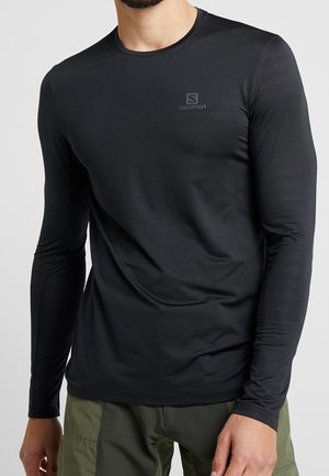 Homme portant un t-shirt Salomon noir ajusté à manches longues et un pantalon cargo vert, debout devant un fond clair uni.
