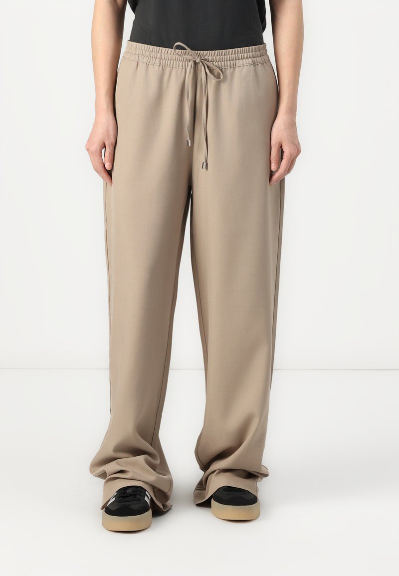 Pantalons beige amples avec une taille élastique et un cordon de serrage. Texture lisse et coutures latérales, associés à des baskets noires.