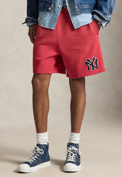 Homme portant un short rouge avec un logo marine, des chaussettes blanches, des baskets montantes bleues et des vestes en jean superposées, les mains dans les poches.