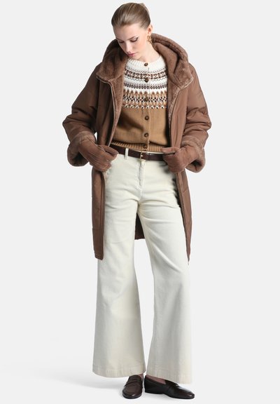 Femme portant un manteau marron doublé de fourrure, un cardigan marron à motifs, un pantalon blanc à jambes larges, des gants marron et des chaussures marron foncé, debout et regardant vers le bas.