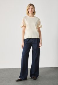 Cream top with crocheted lace sleeves paired with dark blue wide-leg jeans and dark flat shoes, standing on a grey surface.