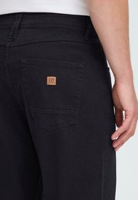 Vyras dėvi juodus kelnes su užpakaline kišenę, kurioje yra mažas rudos spalvos kvadrato logotipo ženklas, ranka ilsisi netoli klubo.
