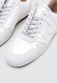Zapatillas blancas con un upper texturizado, que presentan un brillante acento plateado en el collar. Los cordones grises y una suela de goma negra completan el diseño.