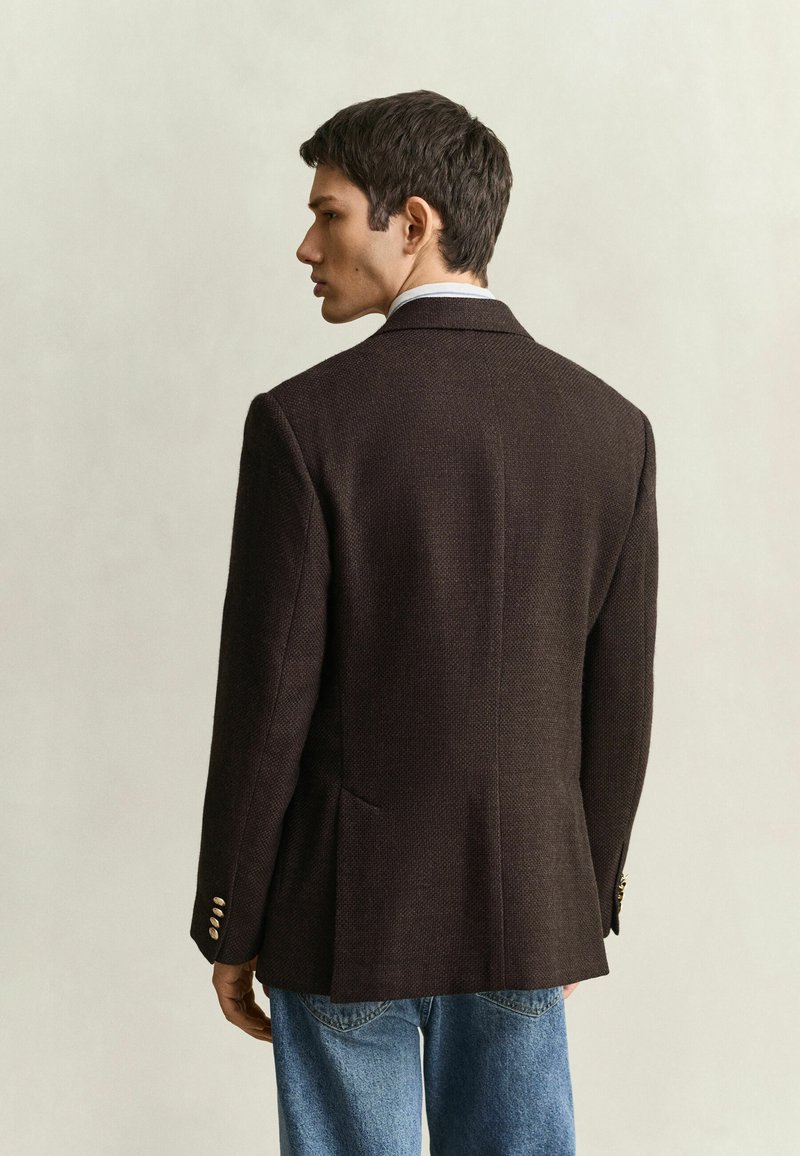 Man standing with back to camera, wearing a dark blazer and blue jeans, facing left against a plain light background.