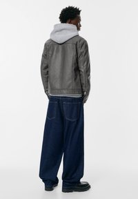 Uomo che indossa un felpa grigia con cappuccio sotto una giacca di pelle grigia, pantaloni jeans scuri a baggy blu e scarpe nere, in piedi rivolto a destra su sfondo bianco.