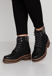 Personne portant des bottines en cuir noir à lacets avec col rembourré gris et semelle en caoutchouc marron, debout sur une surface blanche.