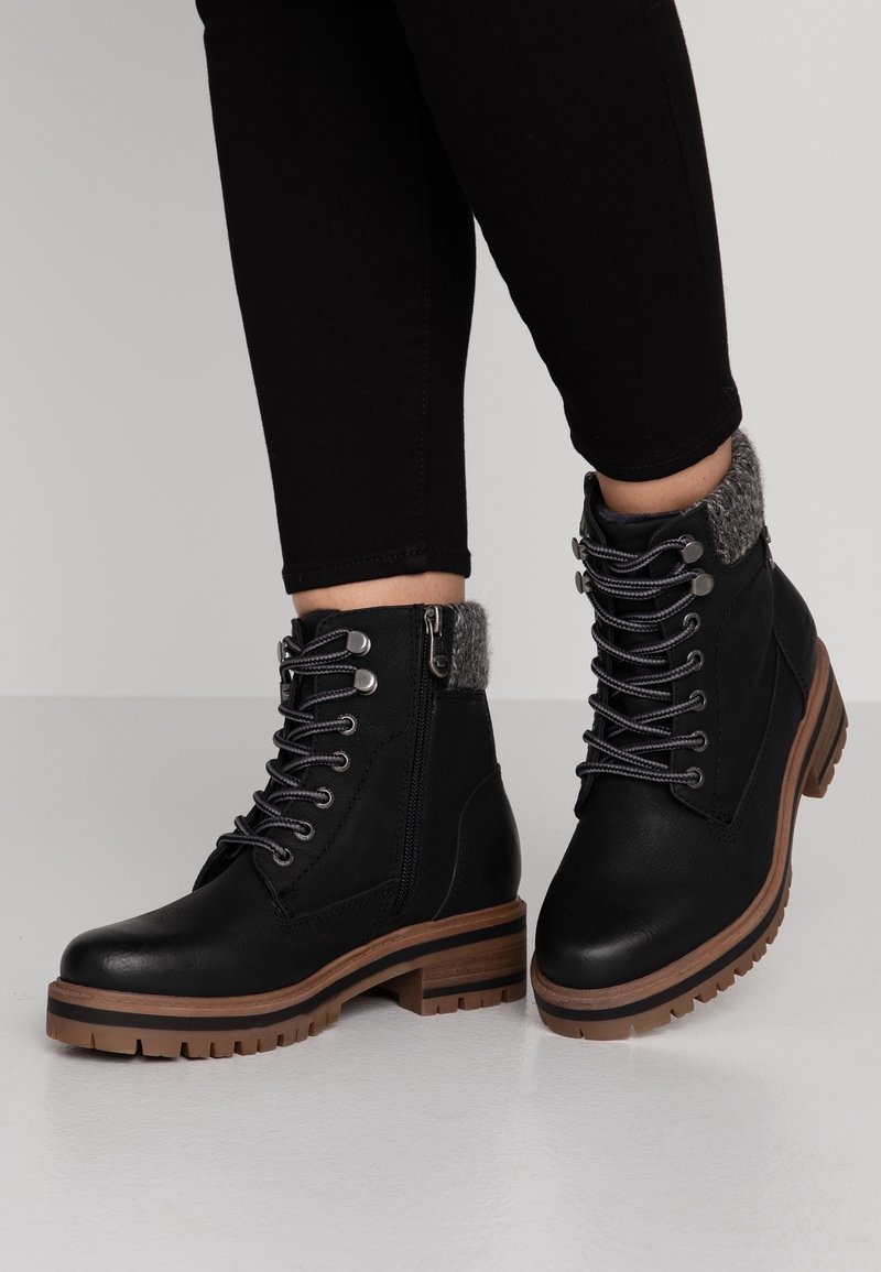 Personne portant des bottines en cuir noir à lacets avec col rembourré gris et semelle en caoutchouc marron, debout sur une surface blanche.