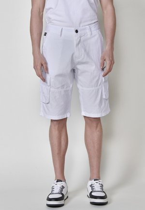 Shorts cargo blancs en tissu léger, dotés de plusieurs poches, d'une fermeture à bouton et d'une coupe droite, associés à des baskets noires et blanches.