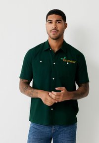 Dunkelgrünes Kurzarm-Hemd mit Knöpfen und zwei Vordertaschen. Mit gesticktem "Guinness"-Logo in Gelb und Grün. Material aus Baumwollmischung.